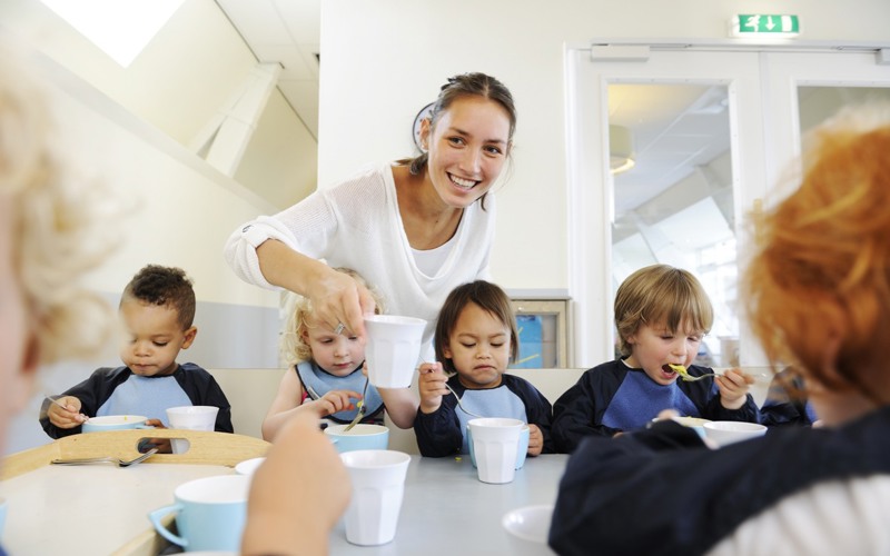 kinderen eten aan tafel en pedagogisch medewerker legt lachend een kopje neer