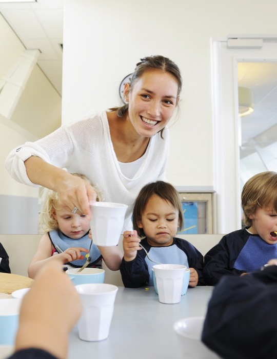 kinderen eten aan tafel en pedagogisch medewerker legt lachend een kopje neer
