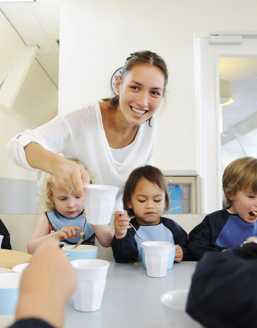 kinderen eten aan tafel en pedagogisch medewerker legt lachend een kopje neer
