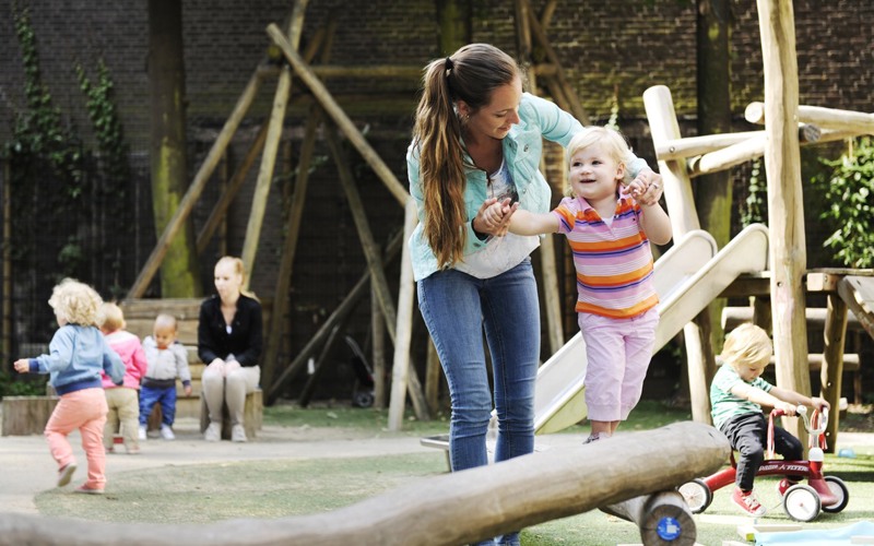 pedagogisch medewerker begeleidt kind op houten balk in speeltuin