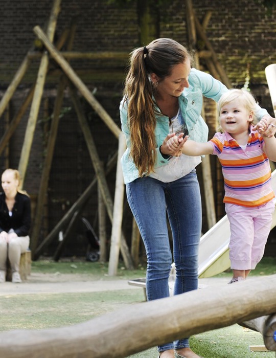 pedagogisch medewerker begeleidt kind op houten balk in speeltuin