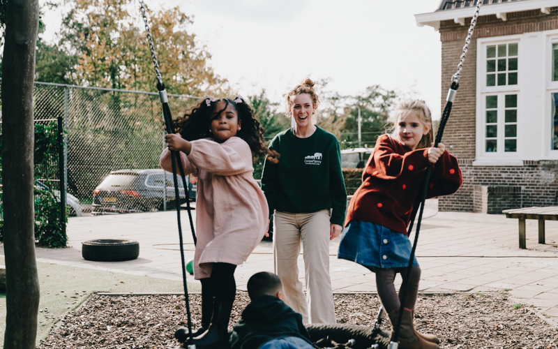 Kinderen op een schommel en een vrouw op de achtergrond die blij toekijkt